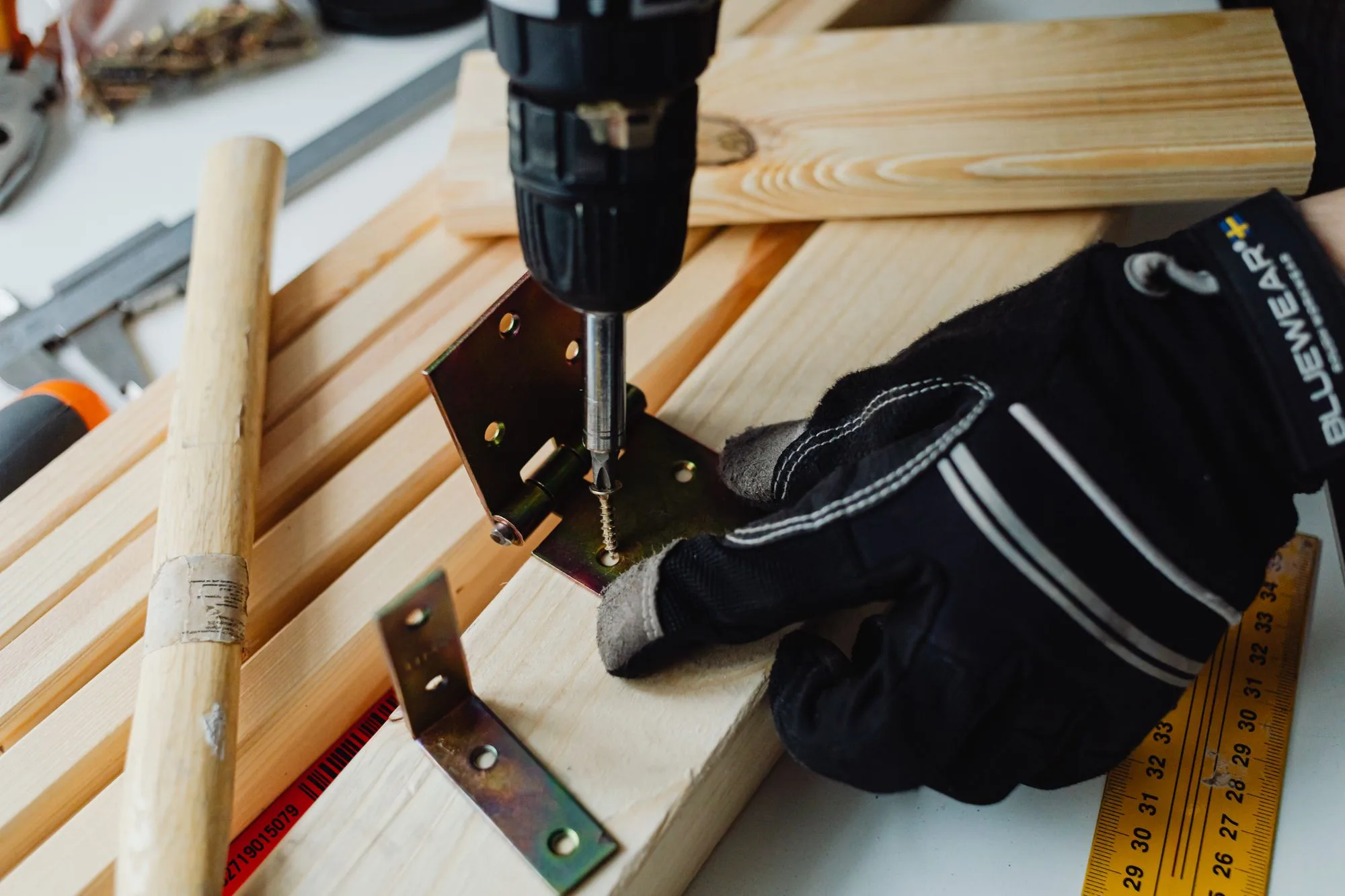 A woodworking project in progress with a power drill, a metal joist hanger bracket, and safety gloves on a workbench