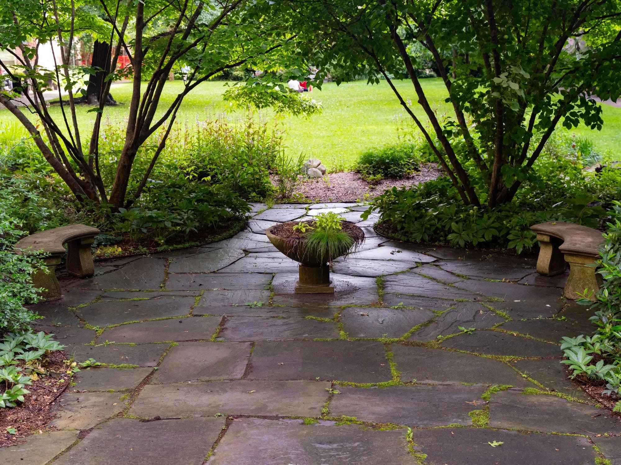 A stone paver patio surrounded by lush greenery with a central raised planting bed