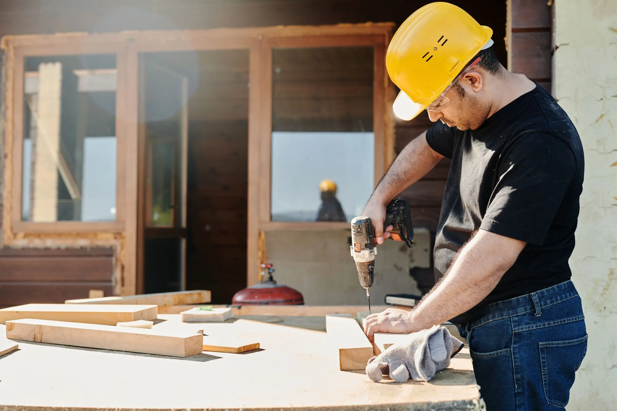 A construction worker in a hard hat using a power drill on a wooden plank at a renovation site